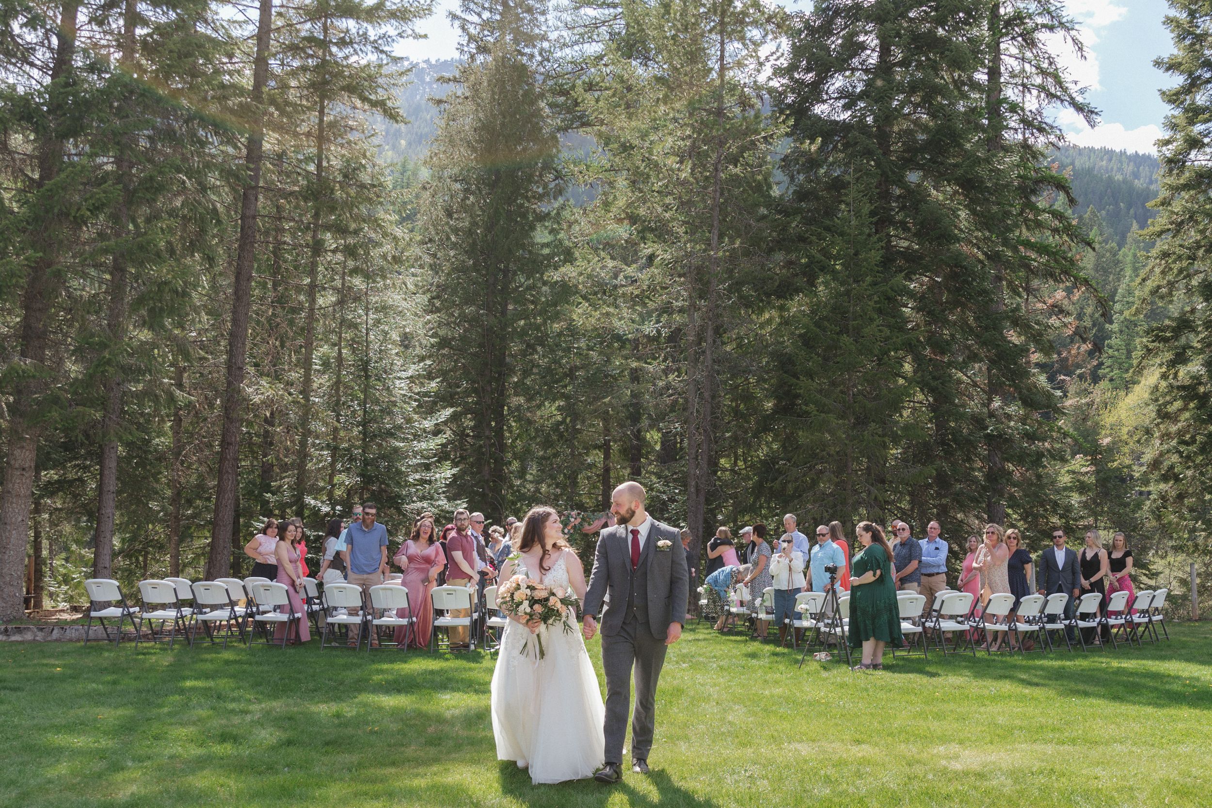 Outdoor wedding ceremony in a scenic forest setting with evergreen trees and guests seated on white chairs on a grass lawn.