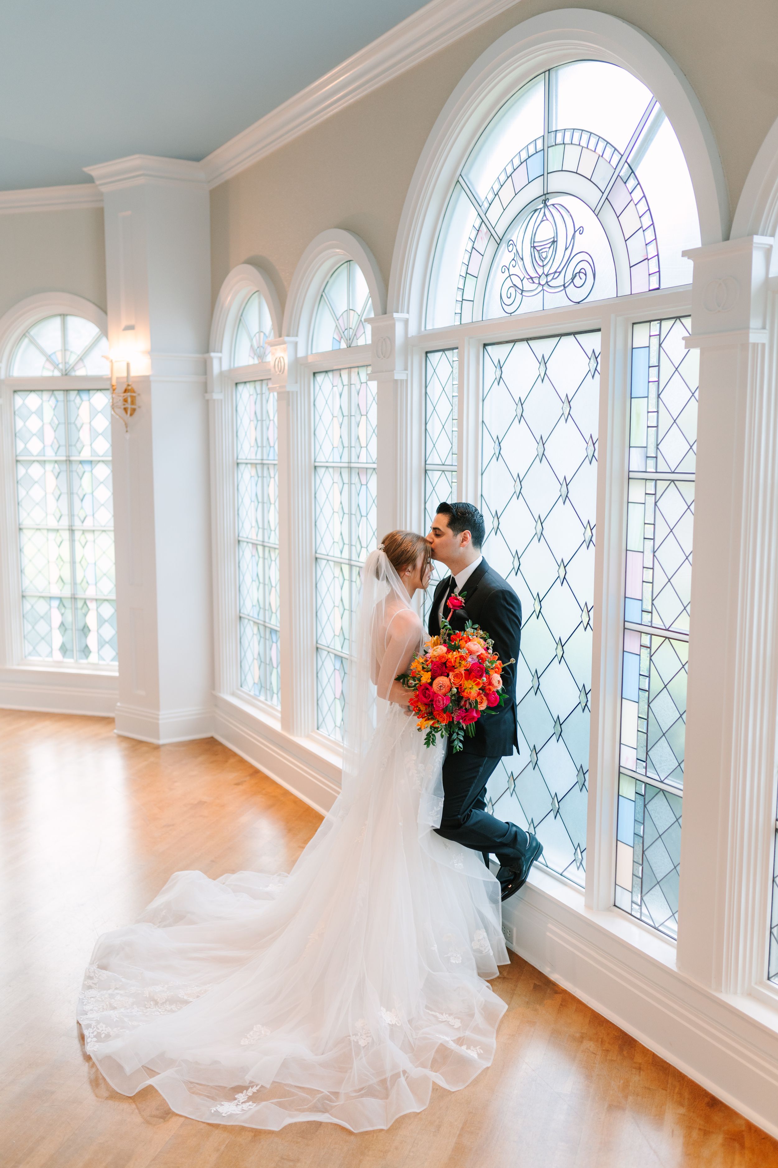 Beautiful arched leaded glass windows illuminate an elegant wedding couple with vibrant orange and red bouquet.