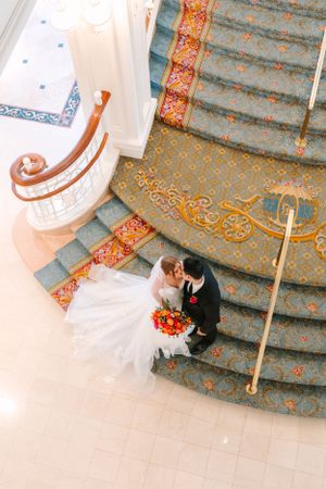 Couple on ornate carpeted staircase with red floral bouquet viewed from above in elegant venue setting.