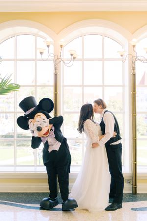 Mickey Mouse mascot poses playfully while a couple shares a romantic moment by tall arched windows at Disney World.