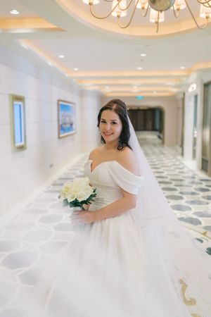A bride in a white wedding dress poses elegantly in a luxurious hallway with ornate ceiling lighting.
