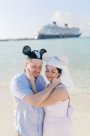 Couple in casual attire embraces on a tropical beach with cruise ships visible in the background.
