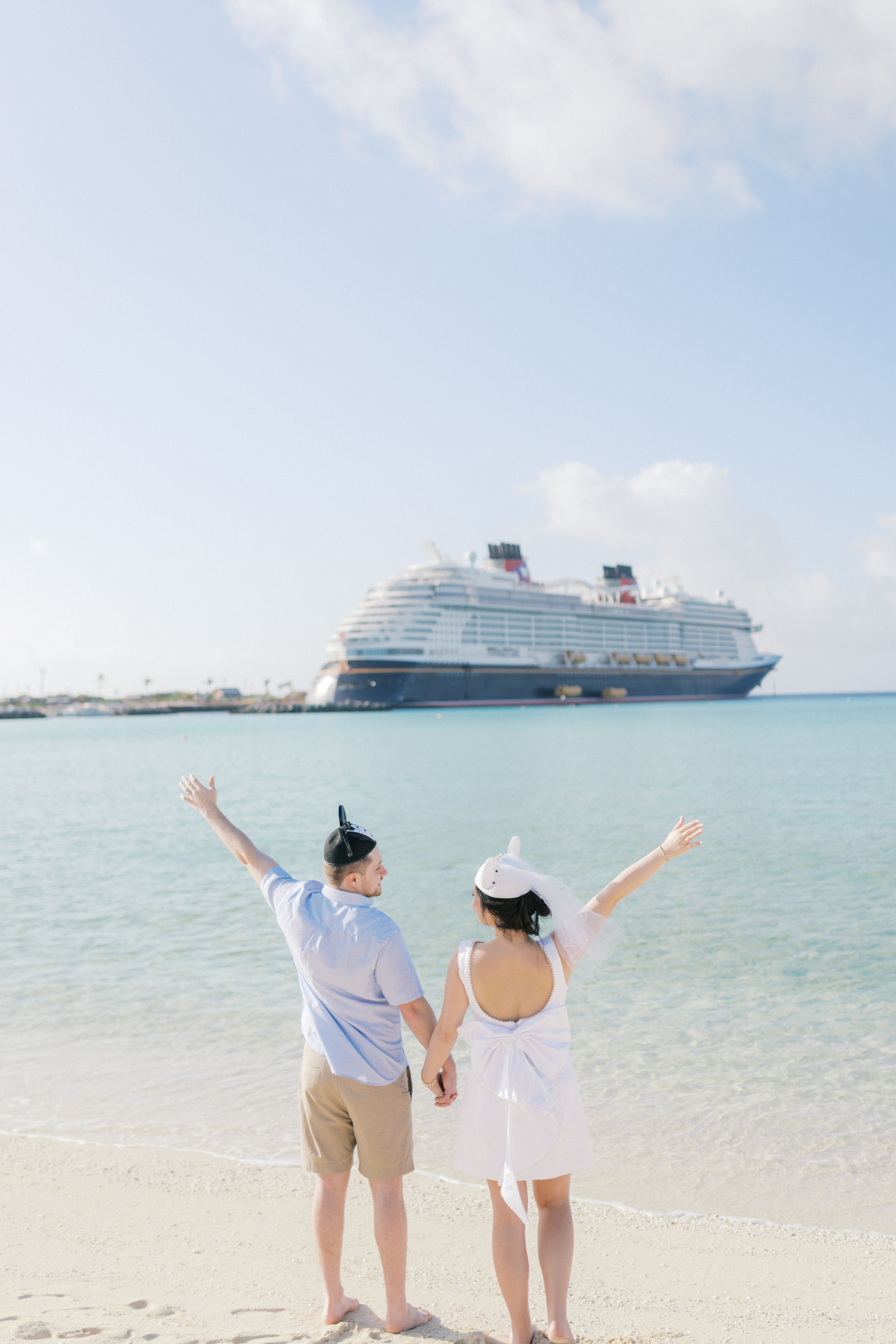 Couple celebrates on a white sandy beach with cruise ships anchored in the turquoise waters behind them.