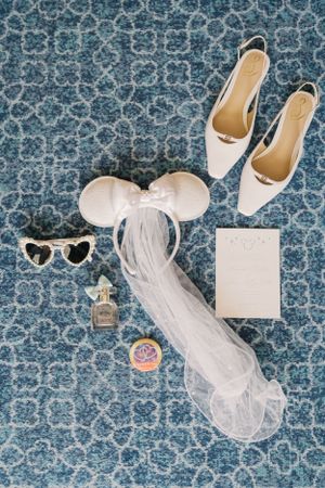 Wedding accessories including a white veil with mouse ears, ballet flats, sunglasses, perfume and card on blue patterned carpet.
