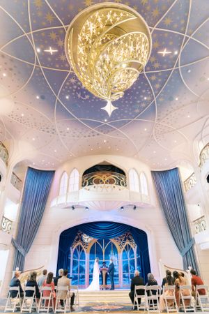Indoor wedding ceremony in an elegant white chapel with blue drapery, ornate chandelier, and Gothic-style architectural details.