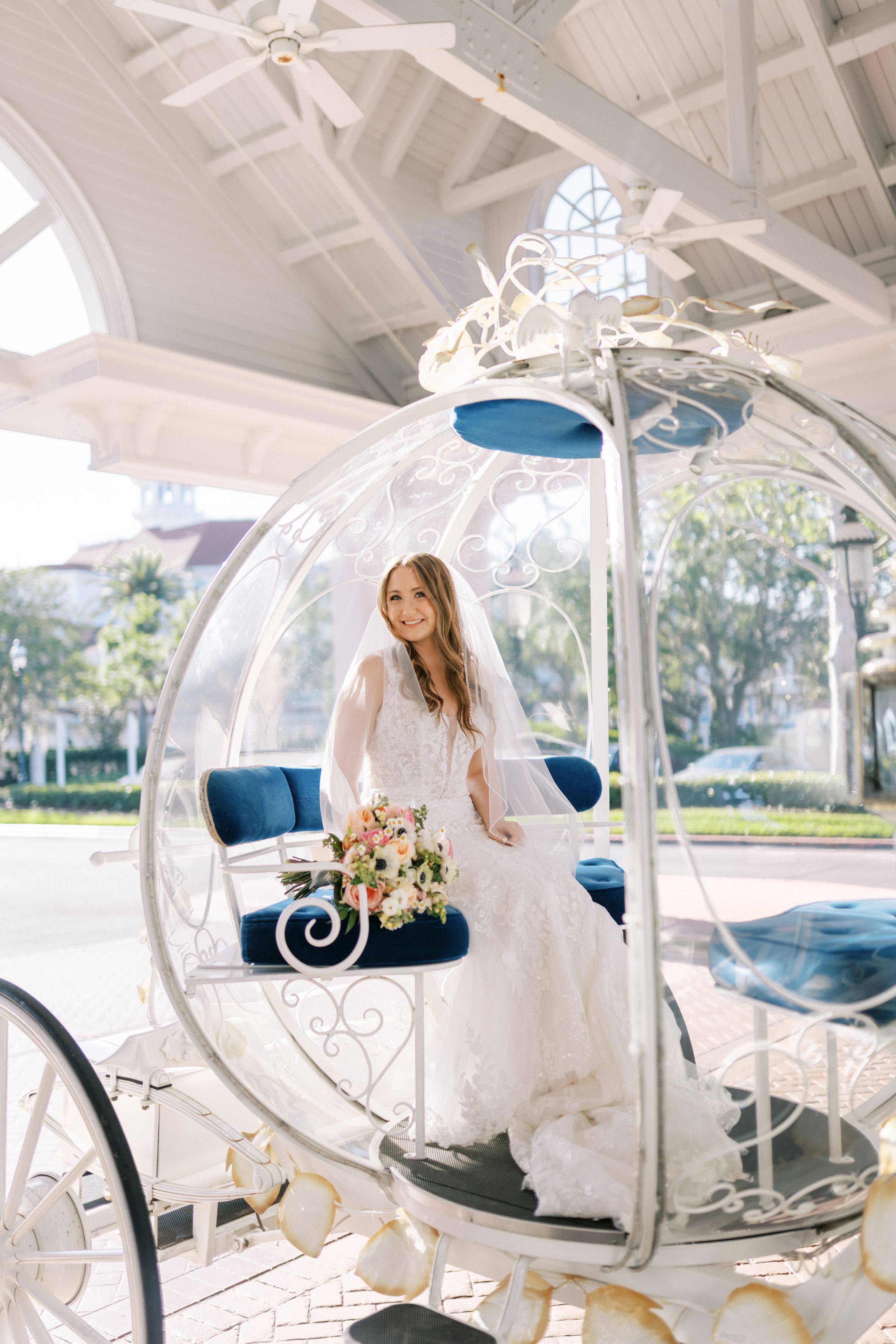 A white ornate horse-drawn carriage with blue velvet seats and floral decorations sits in a bright sunlit venue.
