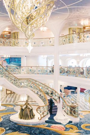 Elegant grand staircase with gold accents and ornate railings aboard a luxury cruise ship features decorative statue and chandelier.