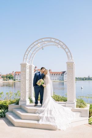 Elegant white wedding arch overlooking a lake with buildings in the background on a sunny day.