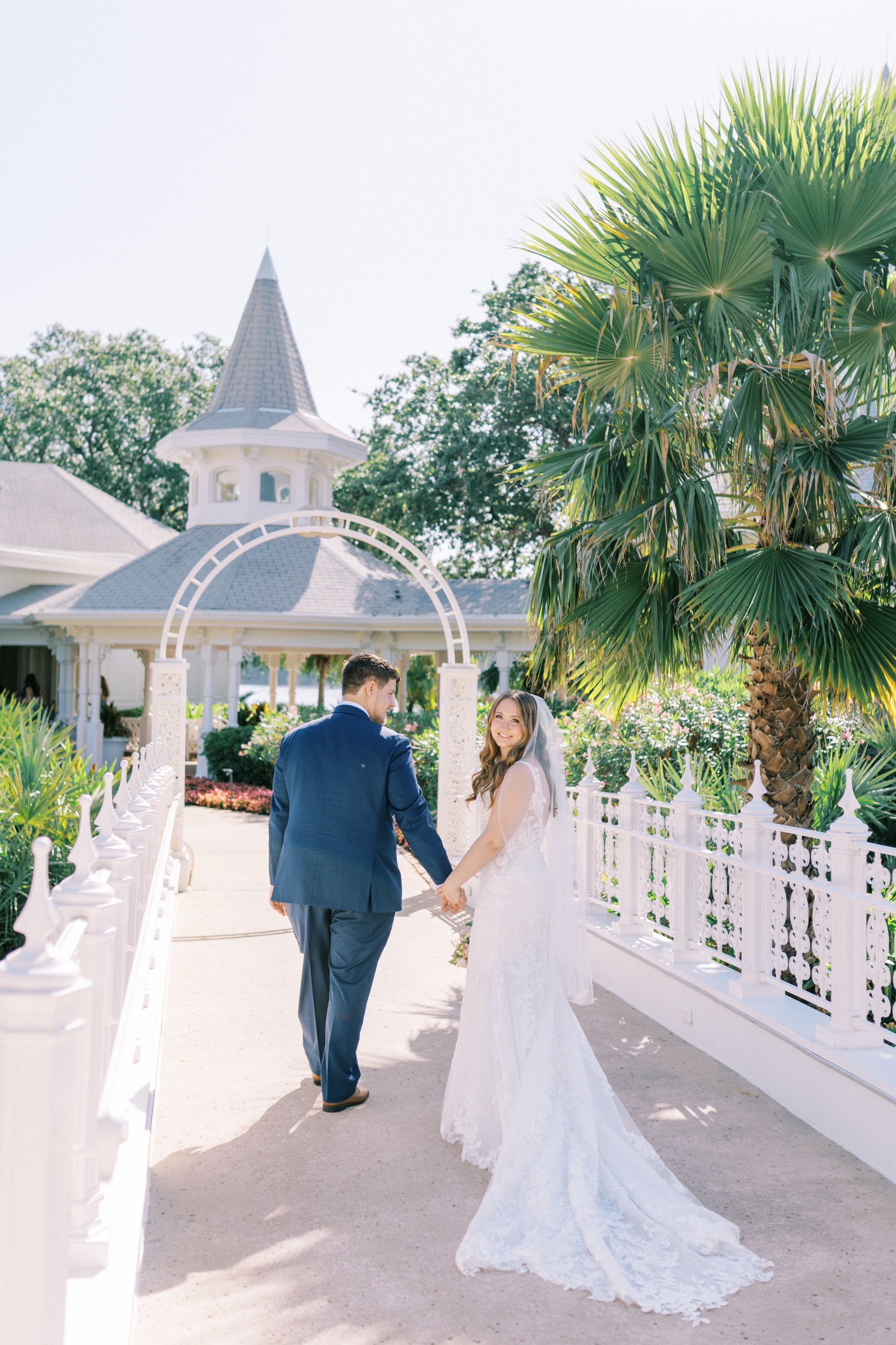 A couple walks along a white-railed walkway at a wedding venue with palm trees and a spired gazebo in the background.