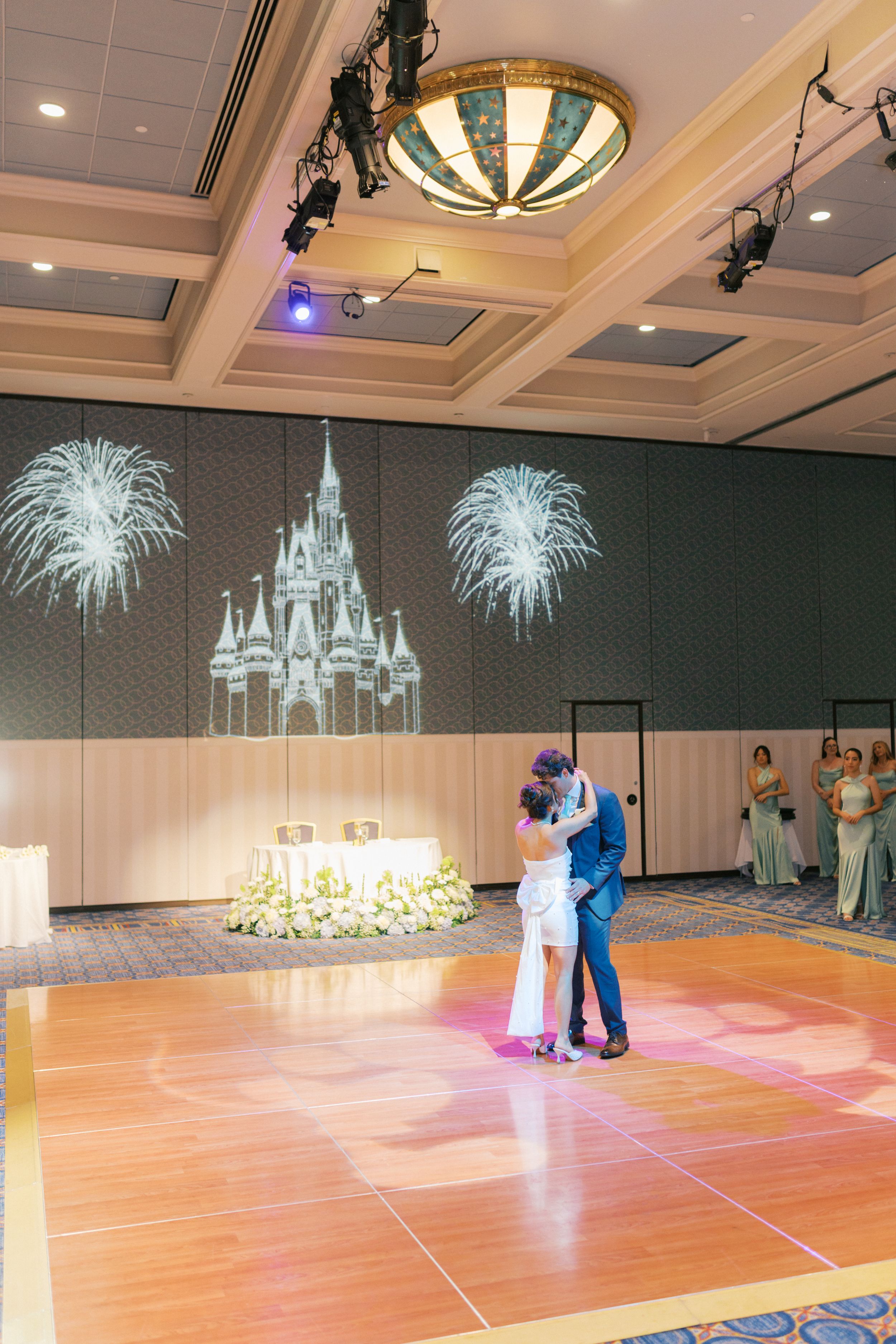 Elegant ballroom with castle-themed chandeliers and projections illuminating a wooden dance floor.