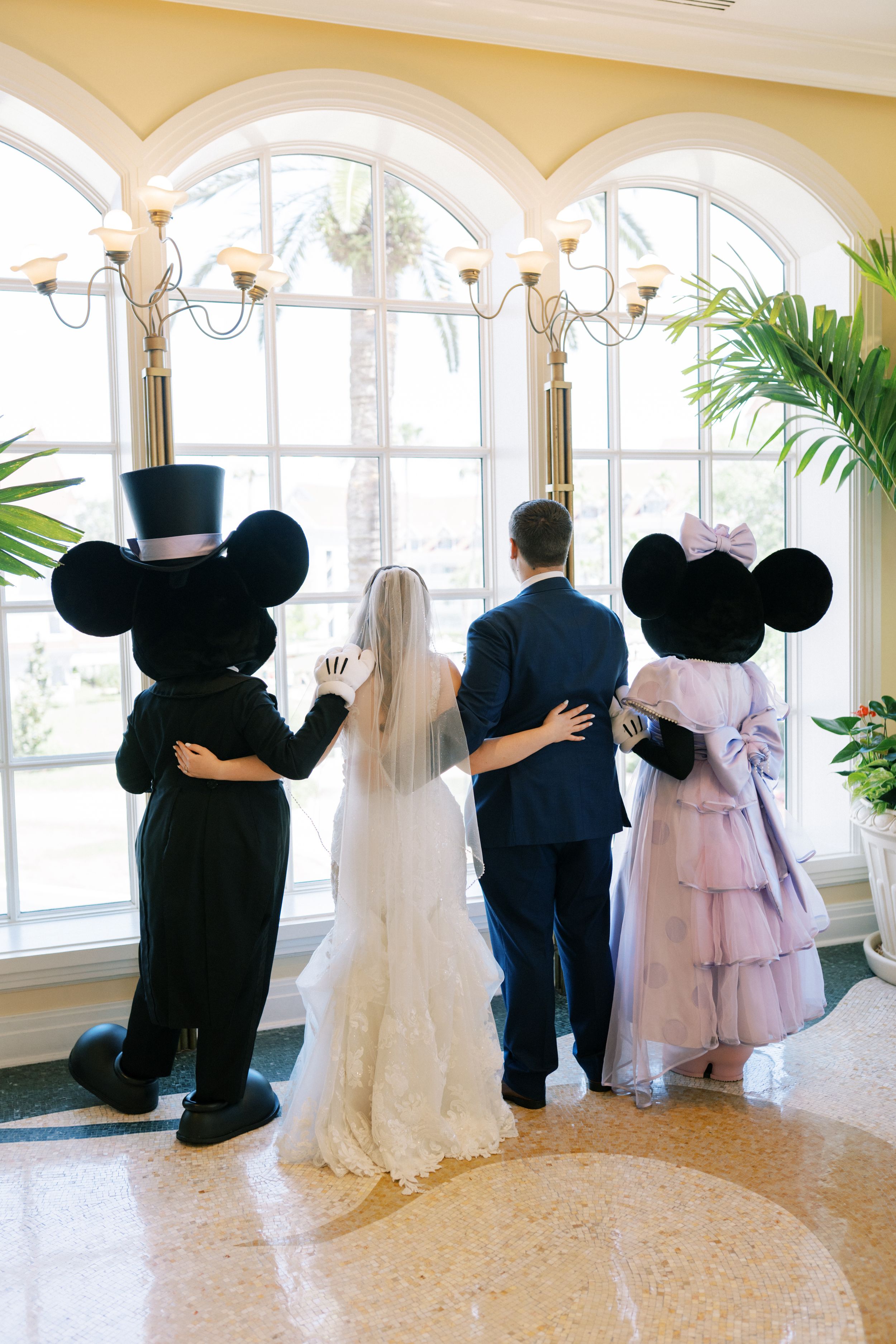 Wedding couple and Disney characters Mickey and Minnie Mouse look out arched windows in elegant venue.