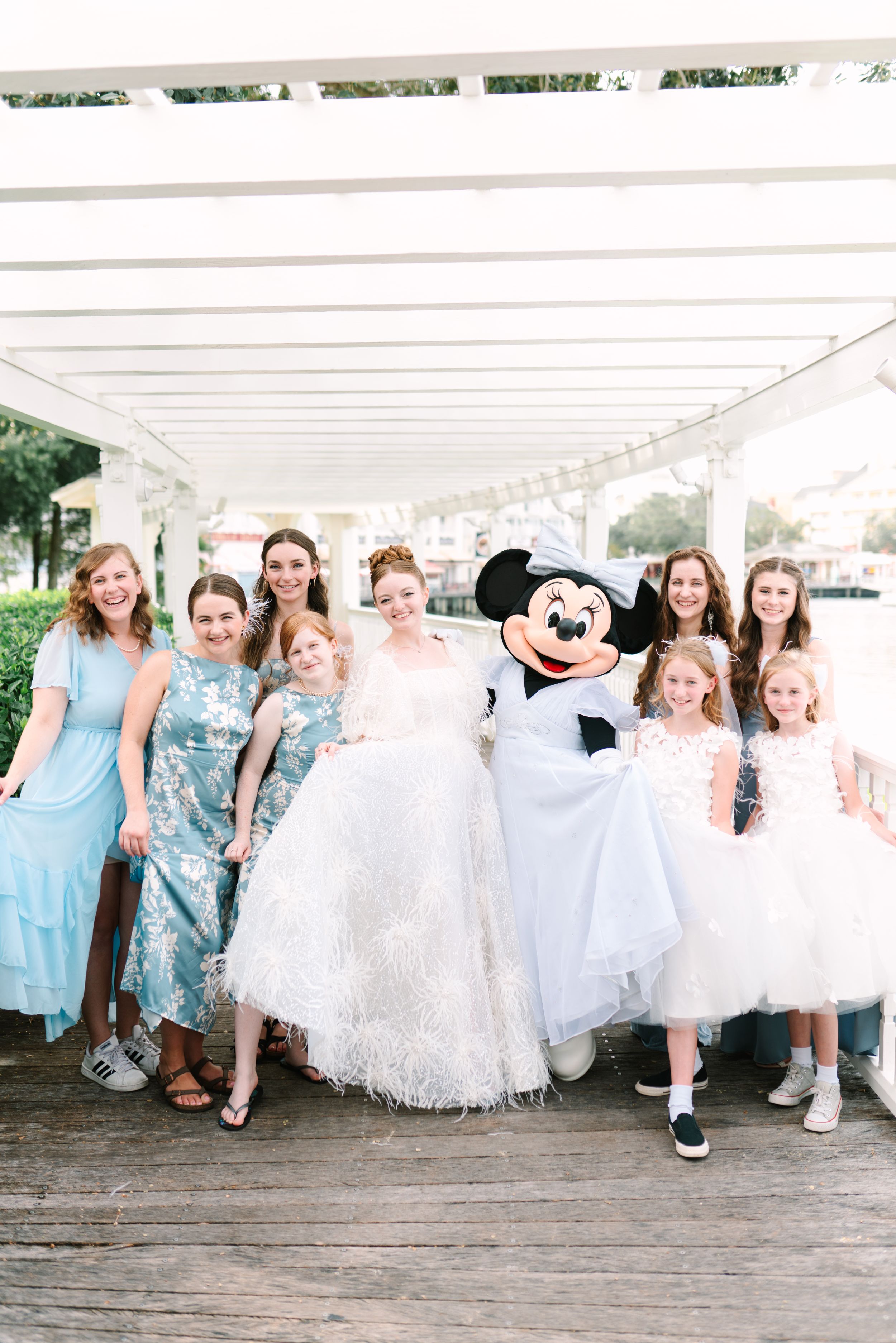 Mickey Mouse character poses with wedding party in white dresses and formal attire on wooden dock.