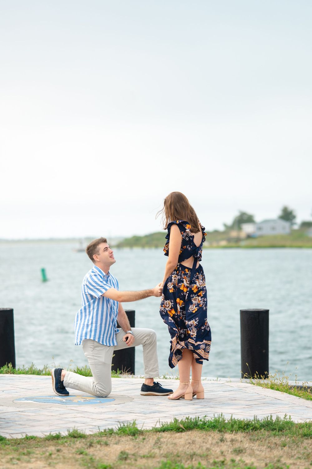 A couple shares a romantic proposal moment on a waterfront dock with water and greenery in the background in Long Island, NY.