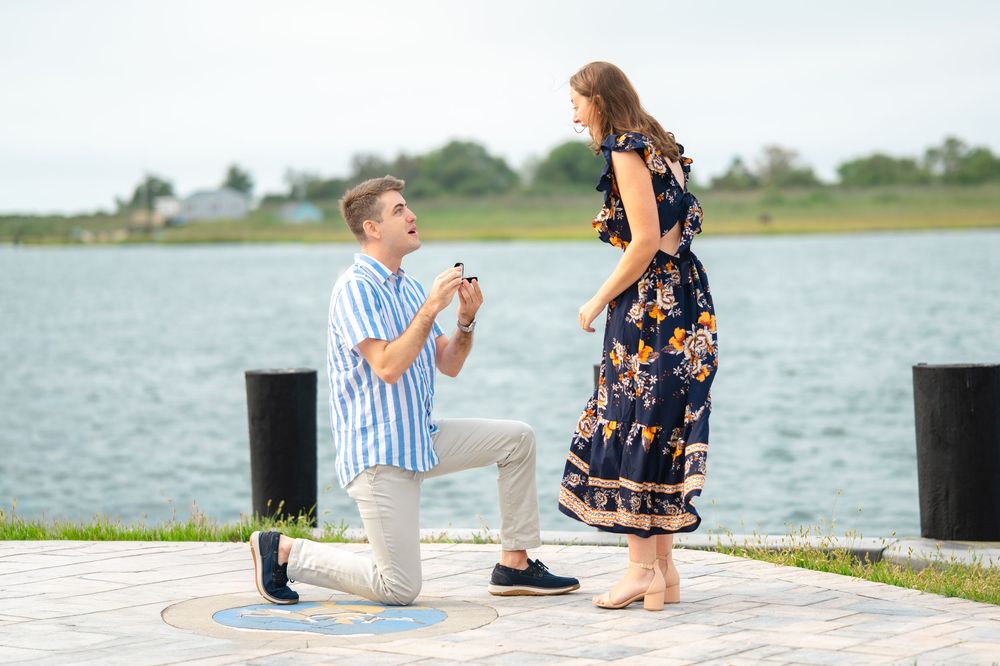 A romantic proposal moment takes place on a wooden dock overlooking a serene waterfront on a sunny day.