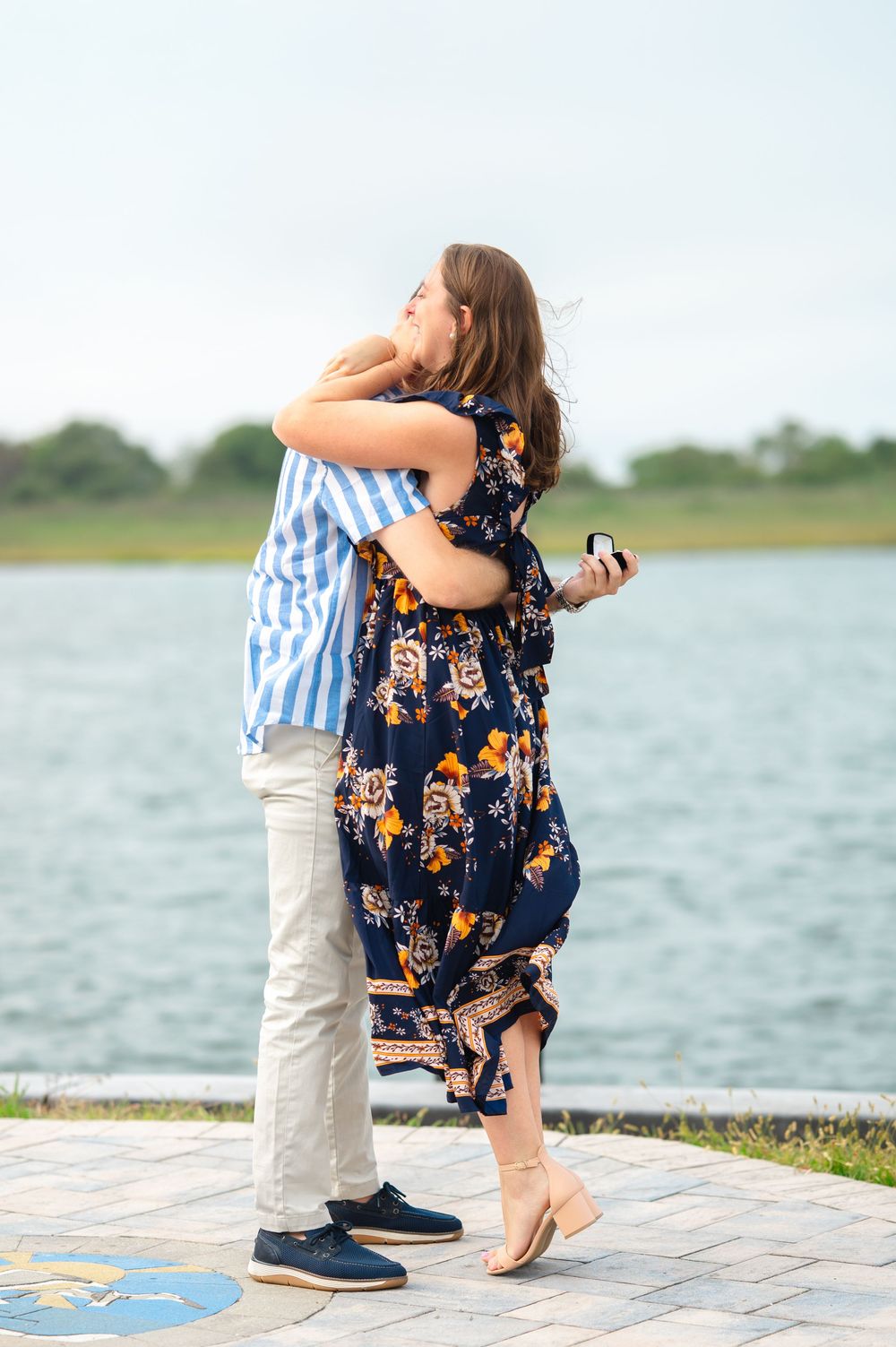 A couple embraces by the waterfront, with her wearing a navy floral dress and him in a striped shirt and khakis.