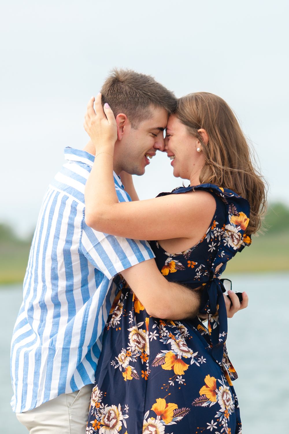 Romantic couple embraces by the water, wearing blue striped shirt and navy floral dress on a sunny day.