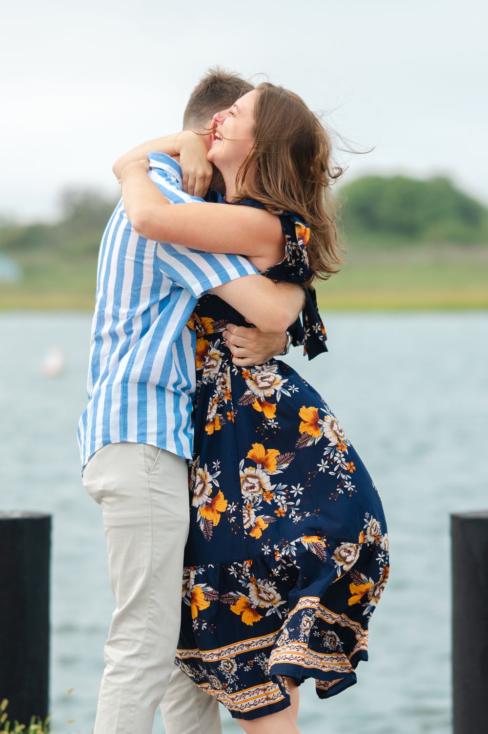Couple embraces by waterfront with woman wearing navy floral dress and man in blue striped shirt and khaki pants.