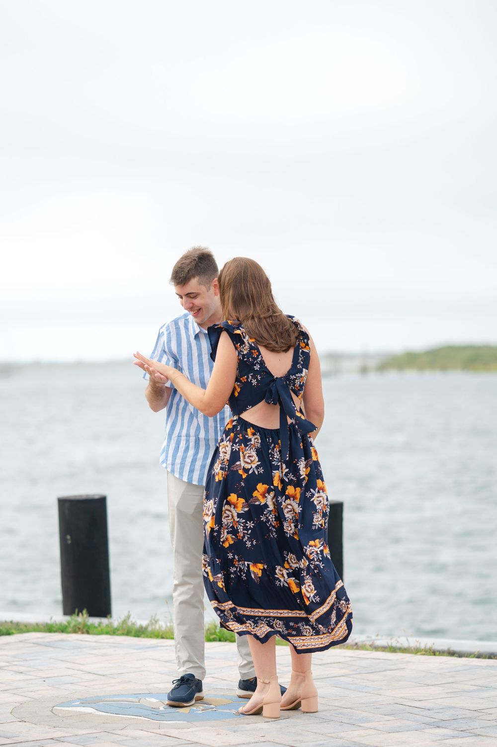 A couple stands together on a waterfront path, dressed in coordinating summer outfits.