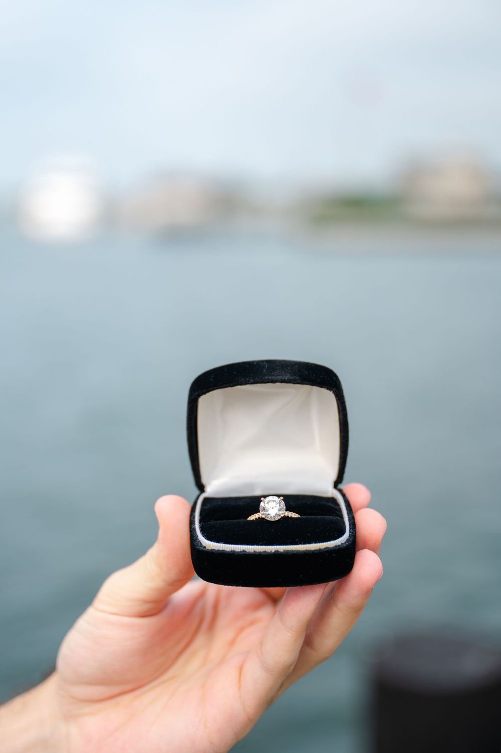 A diamond engagement ring in a black velvet box held up against a blurred waterfront background.