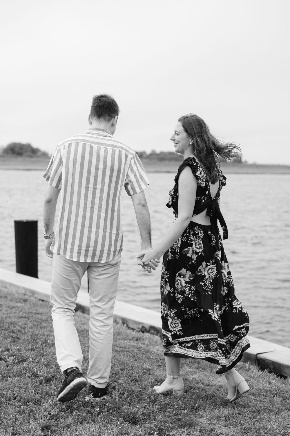 A couple walks hand in hand along a waterfront path in contrasting outfits.