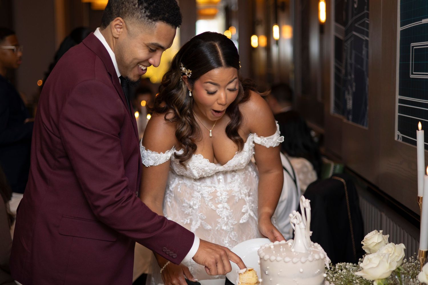 A couple in formal attire cuts their elegant white wedding cake together at an indoor reception.