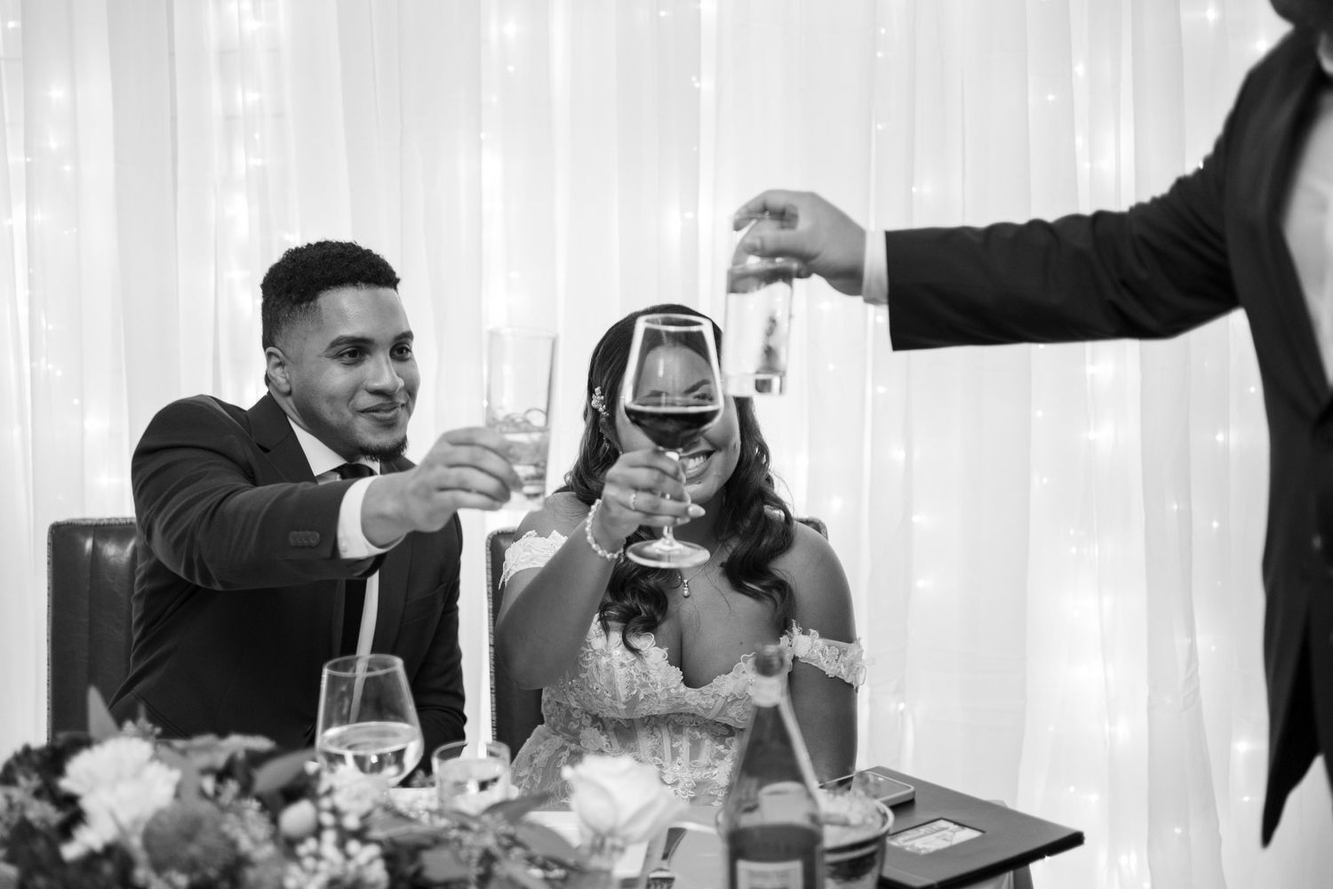 A black and white photo of wedding guests toasting with wine glasses at a reception celebration.