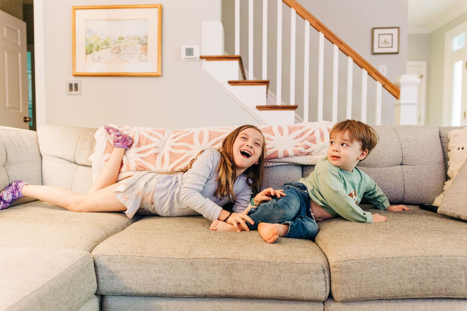 Young siblings lounging and laughing together on a comfortable gray couch in a bright living room with white walls and stairs.