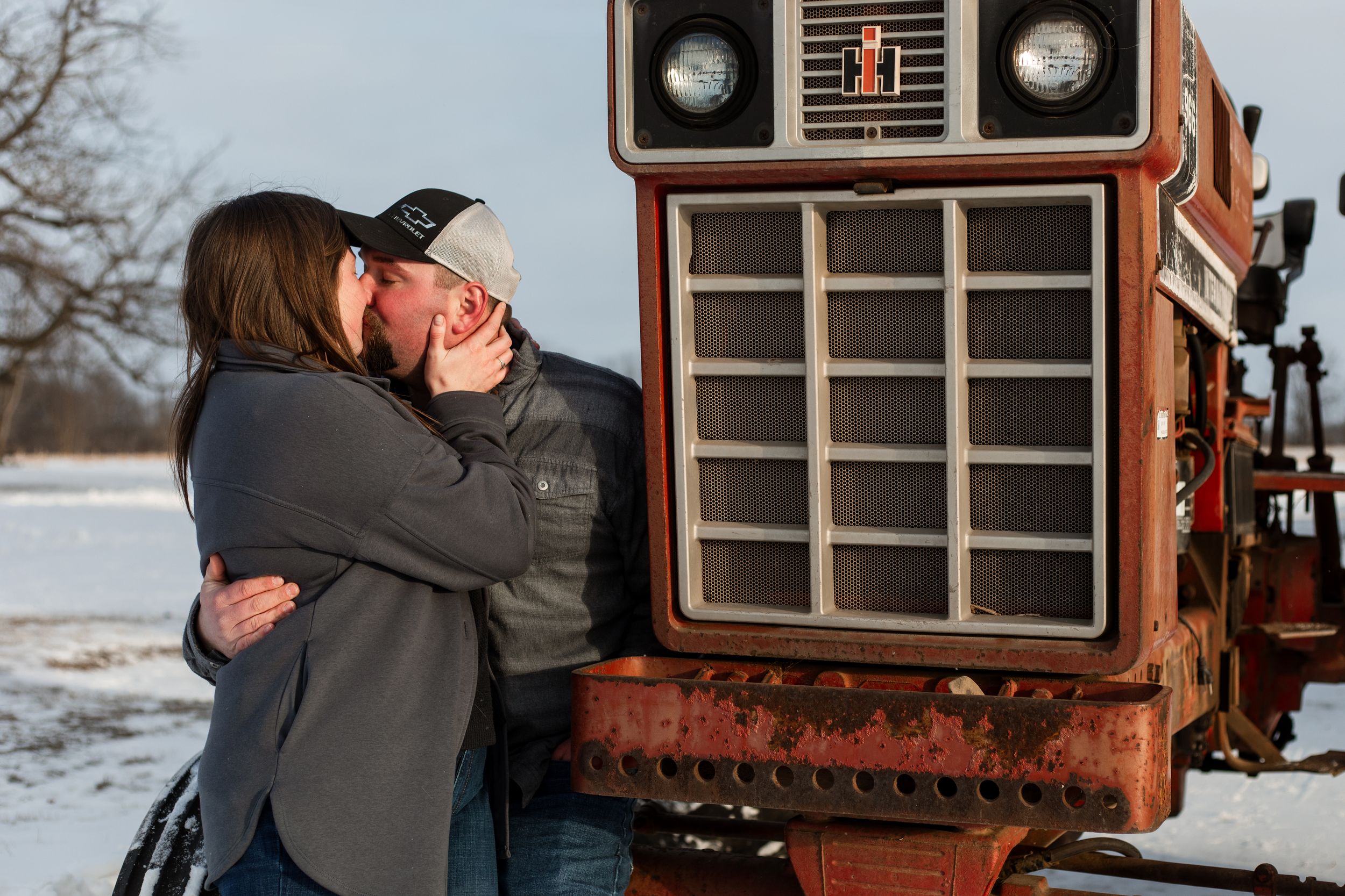 A couple embraces in front of a vintage orange tractor in a snowy winter landscape during golden hour.