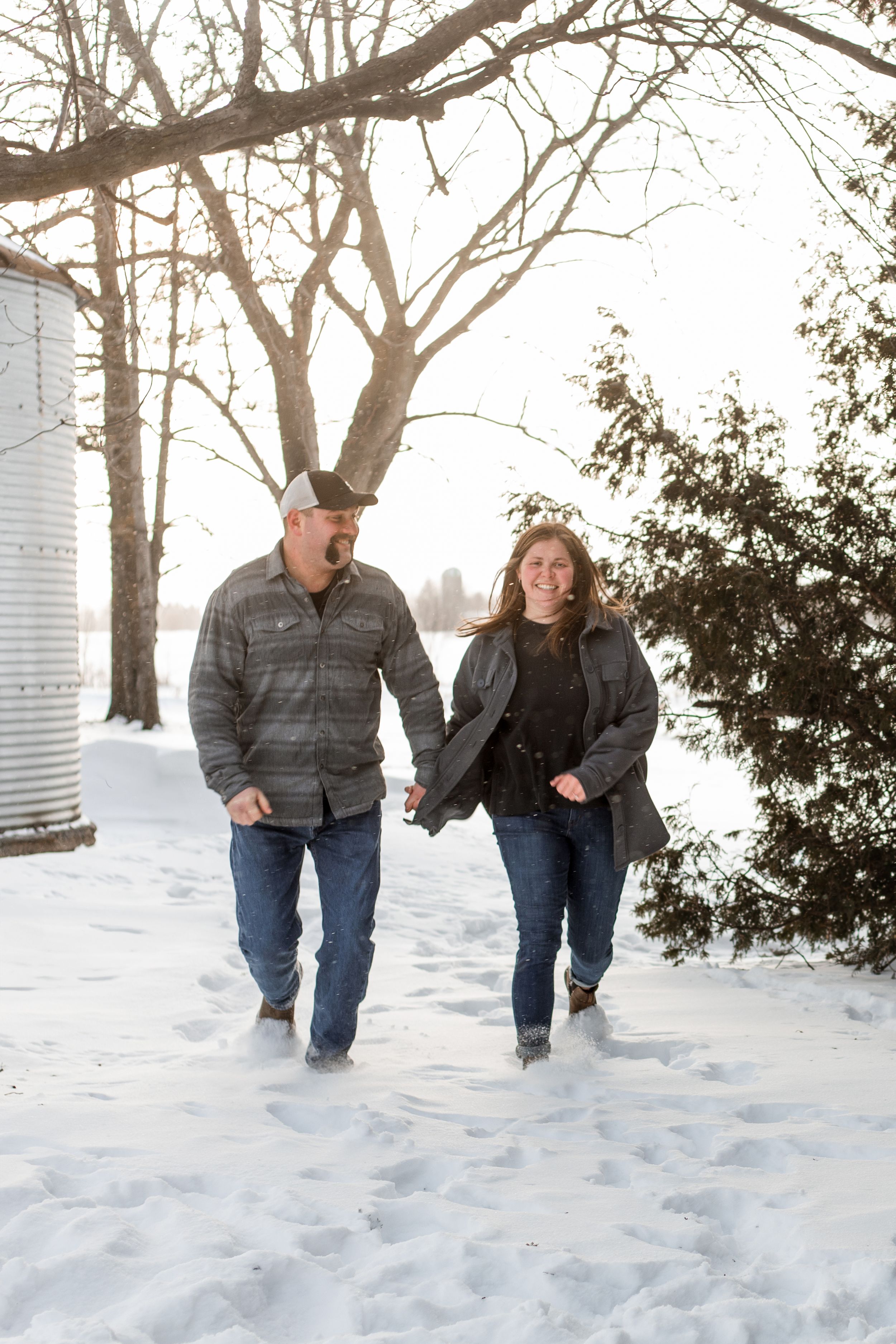 A couple walks together holding hands in a snowy winter landscape with bare trees and evergreens in the background.
