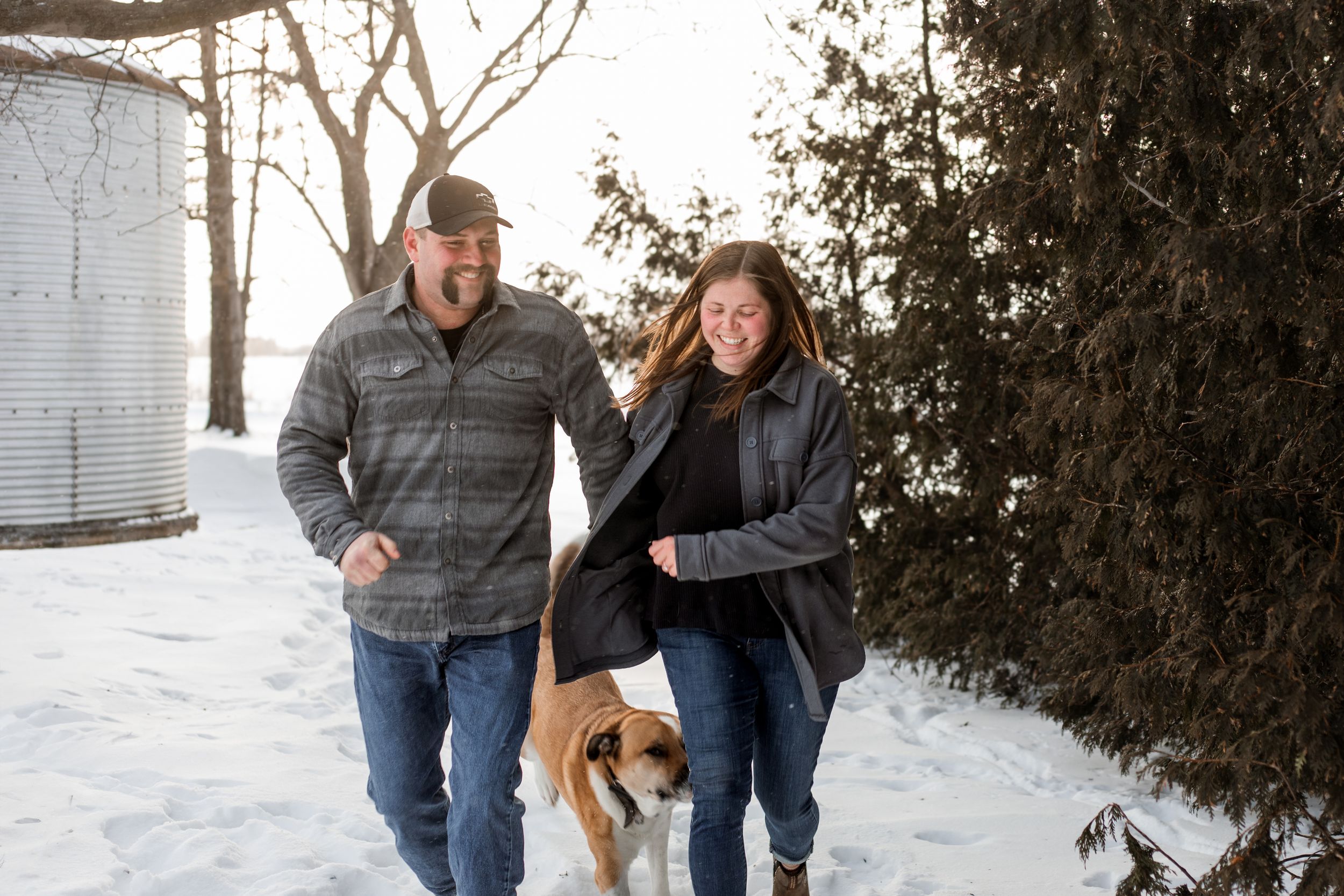 Two people walk together through a snowy winter landscape with evergreen trees in the background.