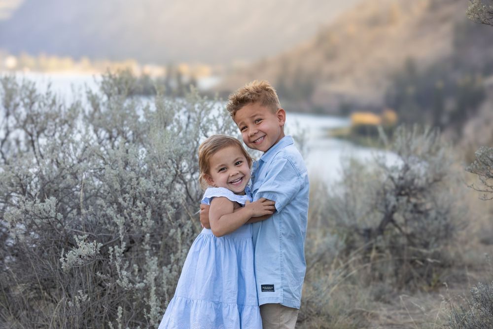 Young brother and sister hugging and smiling in a scenic lakeside shrub landscape.