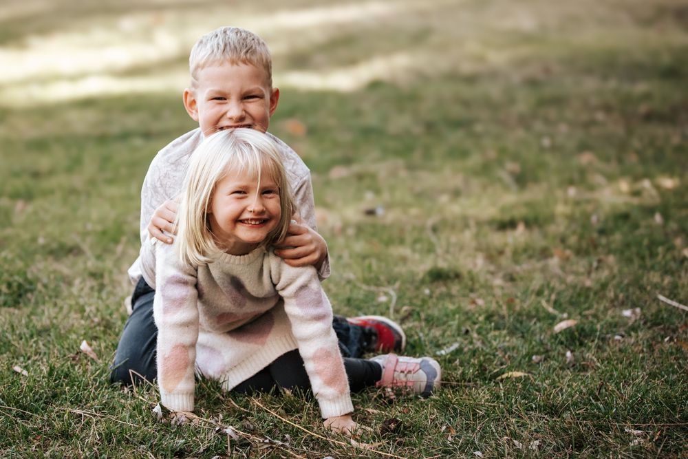 Two blonde siblings hugging and smiling while sitting on green grass outdoors.