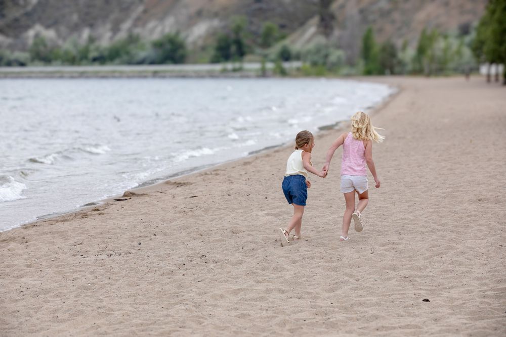Two children running hand in hand along a sandy beach near the water.