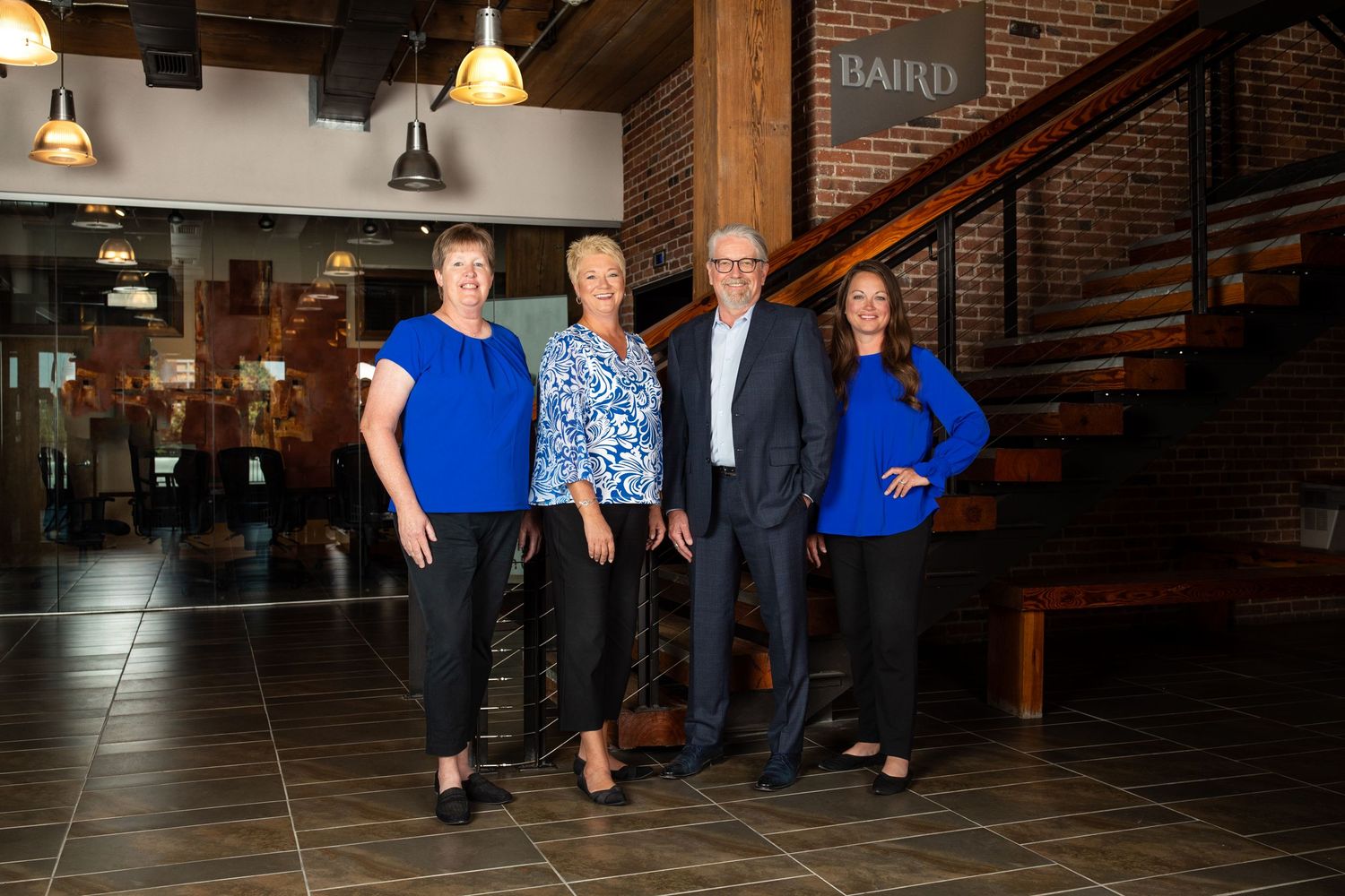 A group of four professionals in coordinated blue and black business attire stand together in a modern brick-walled lobby for a team photo.