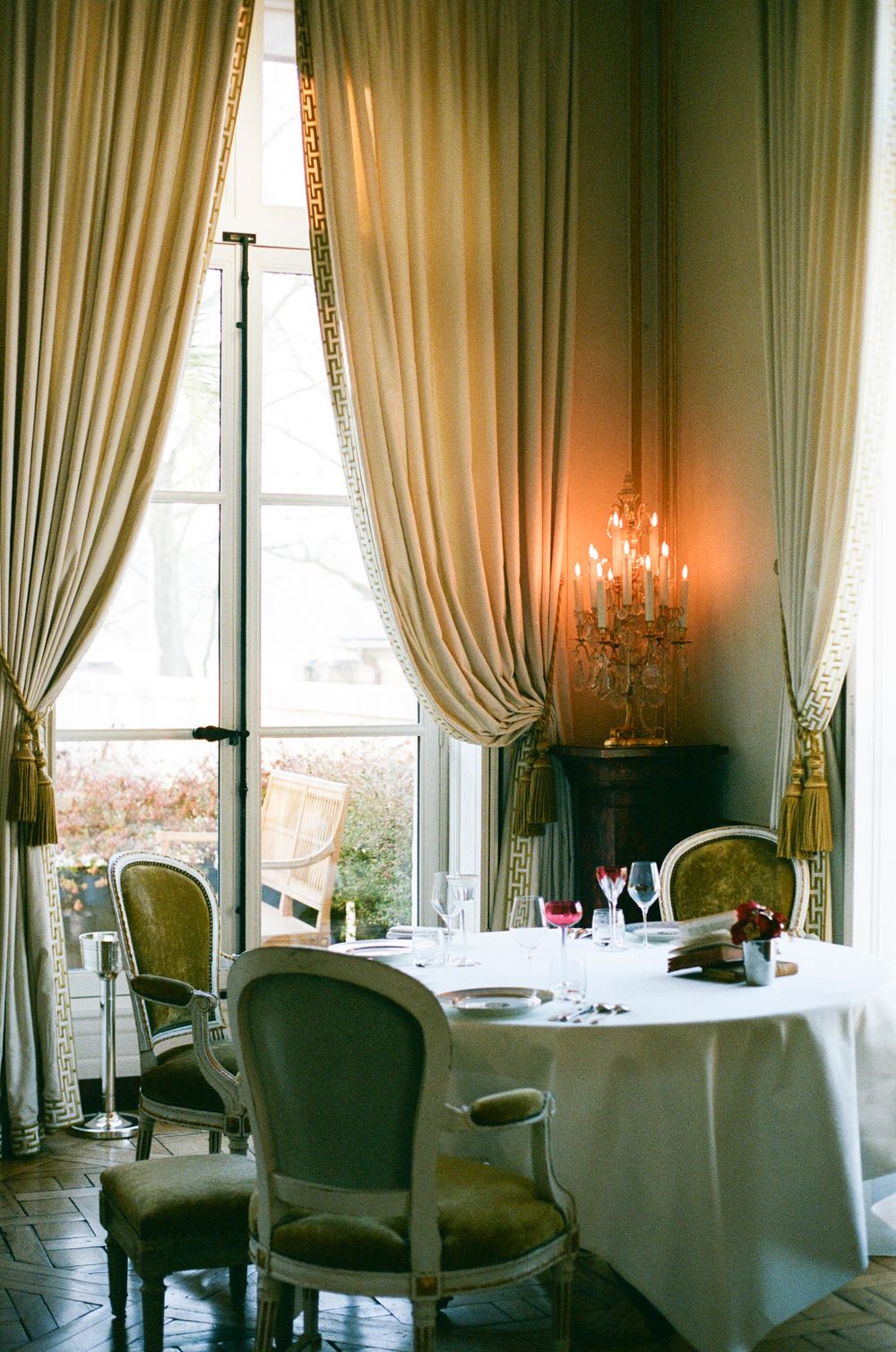 Elegant restaurant interior with tall windows, white tablecloths, and classic French-style chairs in soft lighting. Paris film photography.