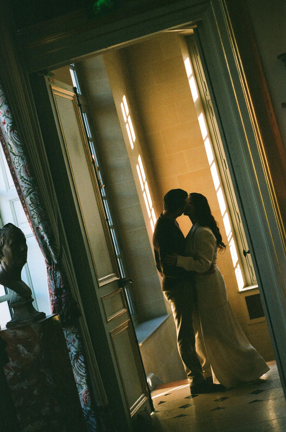 A silhouetted couple embraces in a doorway with dramatic window light streaming in from tall windows. Paris film photographer