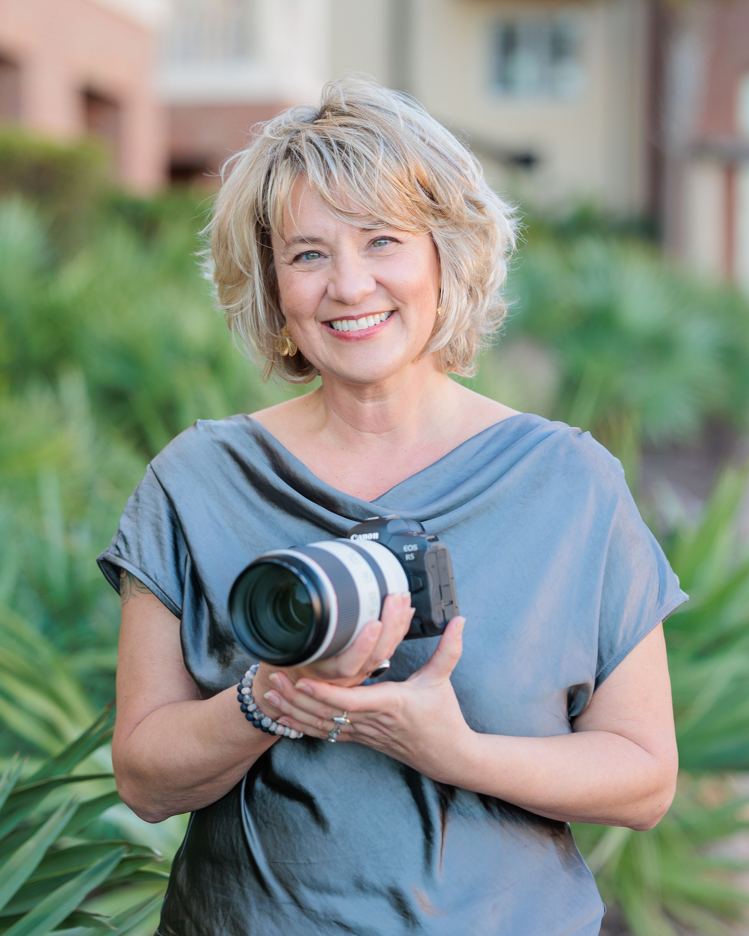 Photographer Amy Stevens in gray blouse holds camera equipment in outdoor garden setting with brick building in background.