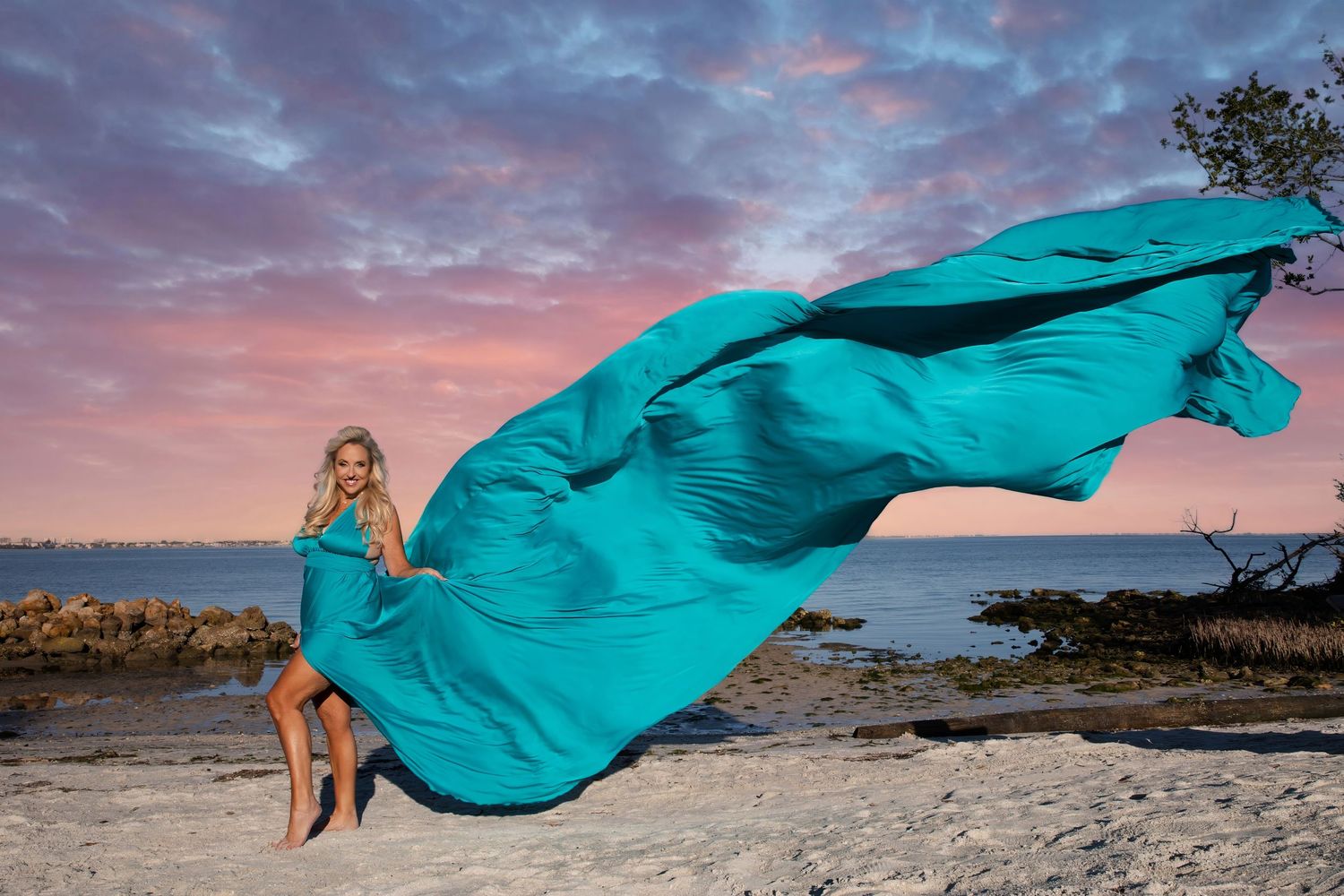 A flowing teal fabric dances in the wind against a dramatic sunset sky during a beach photoshoot. Flying Dress Pictures