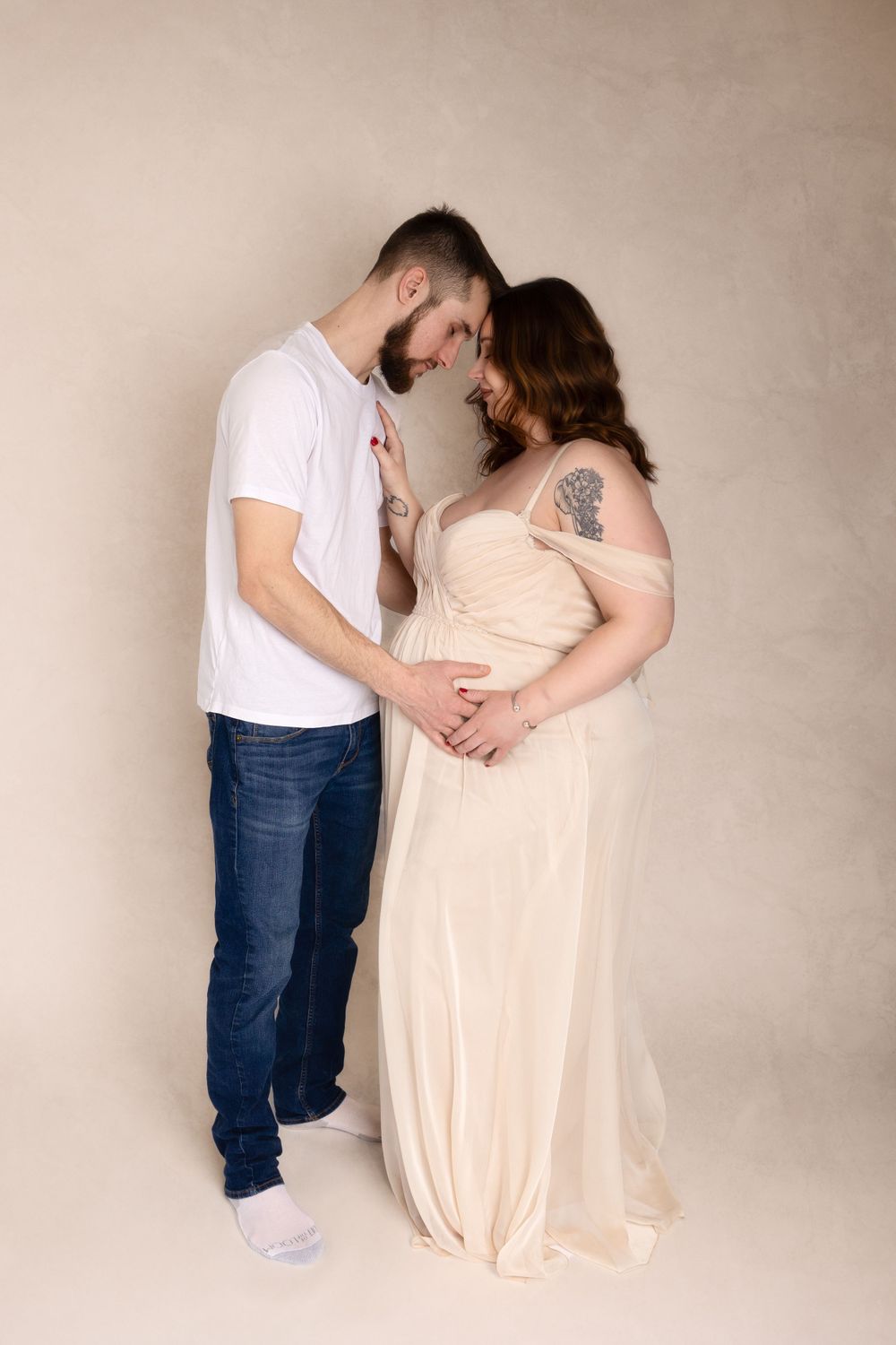 Intimate maternity portrait with couple in cream gown and casual attire against beige background.