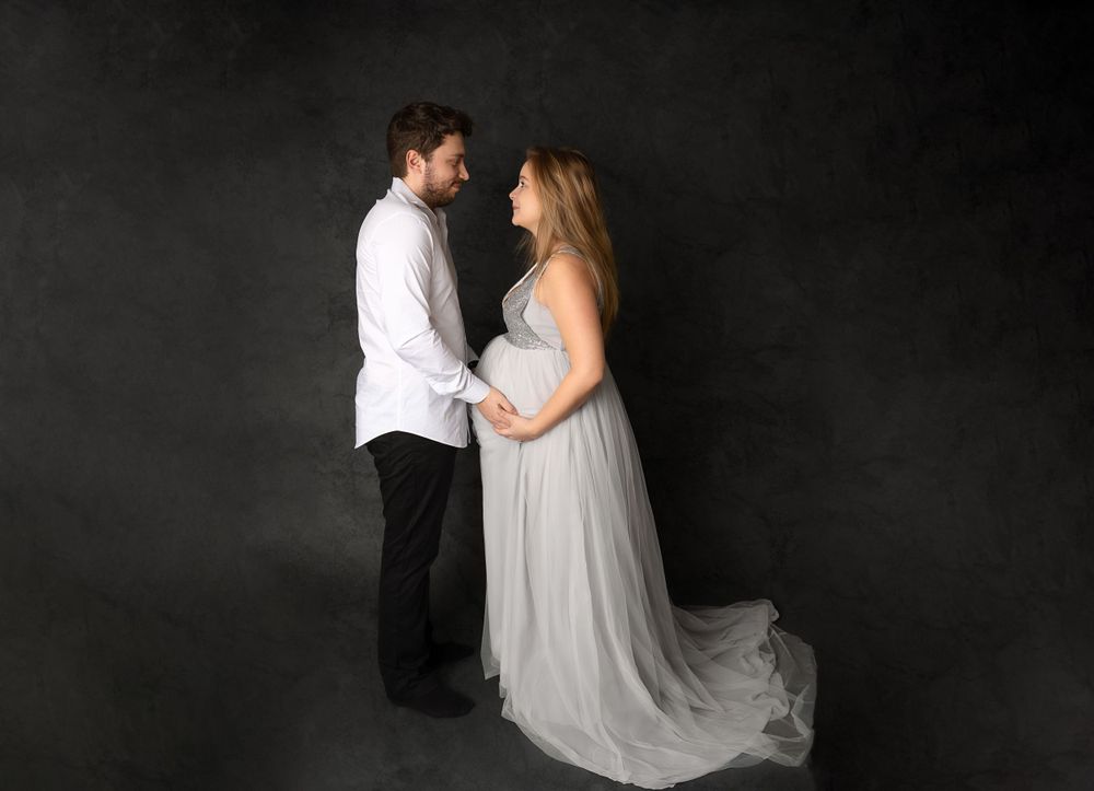 Romantic maternity silhouette of couple in white and grey attire against dark backdrop.