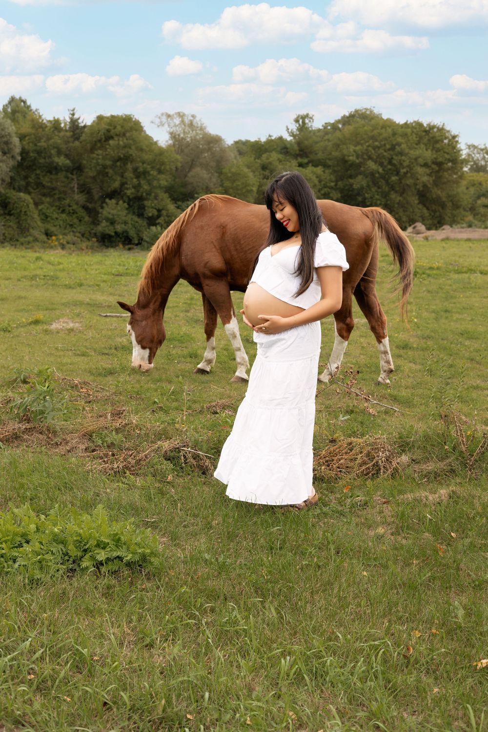 A person in a flowing white dress stands in a grassy field while a brown horse grazes in the background.