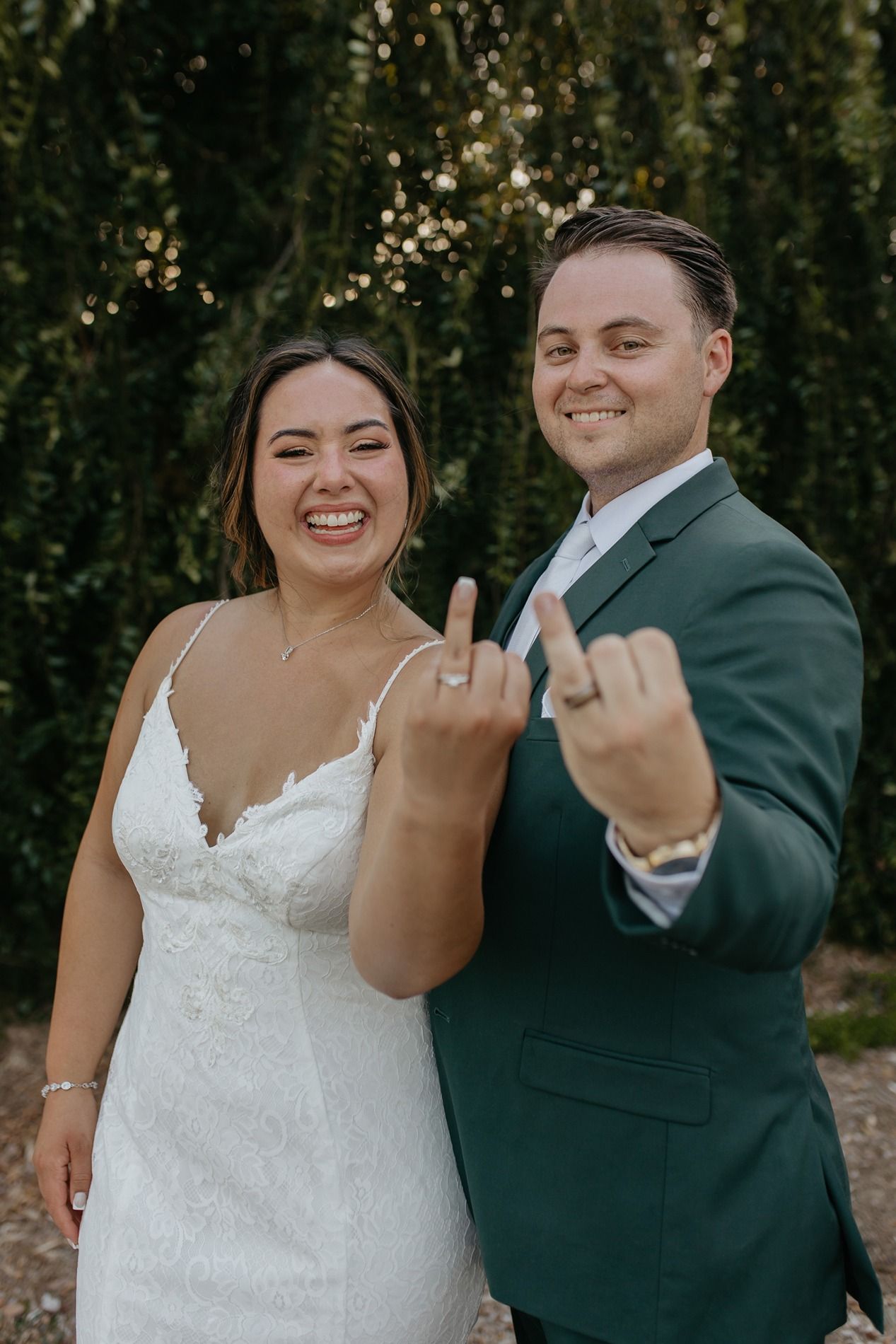 Newlywed couple in wedding attire showing off their rings with playful middle finger gestures in an outdoor setting at Canopy Grove.