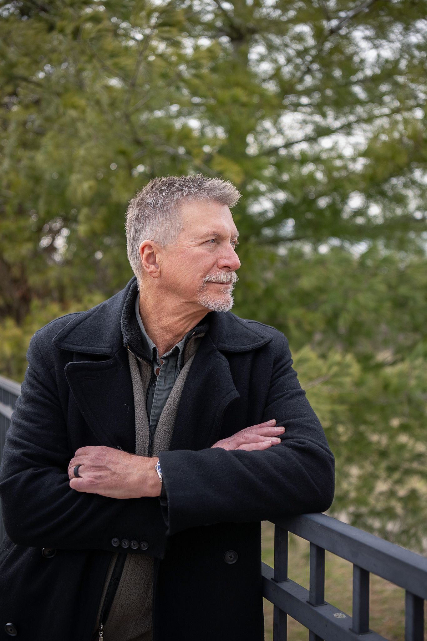 A casually dressed person in a black coat leans against an iron railing with autumn foliage in the background.