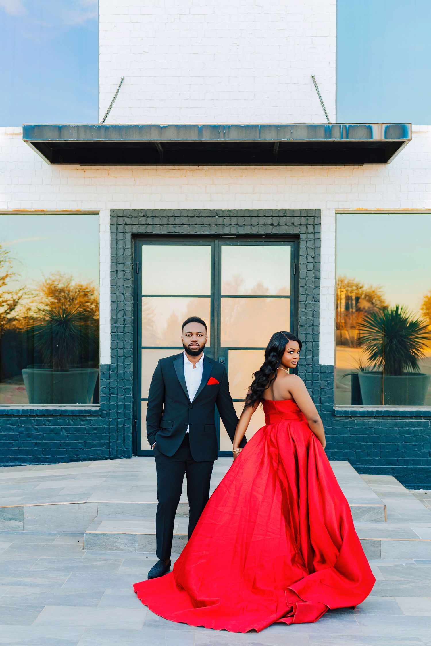 A couple poses in front of a modern building with the woman wearing a flowing red ball gown in a sunset photoshoot.