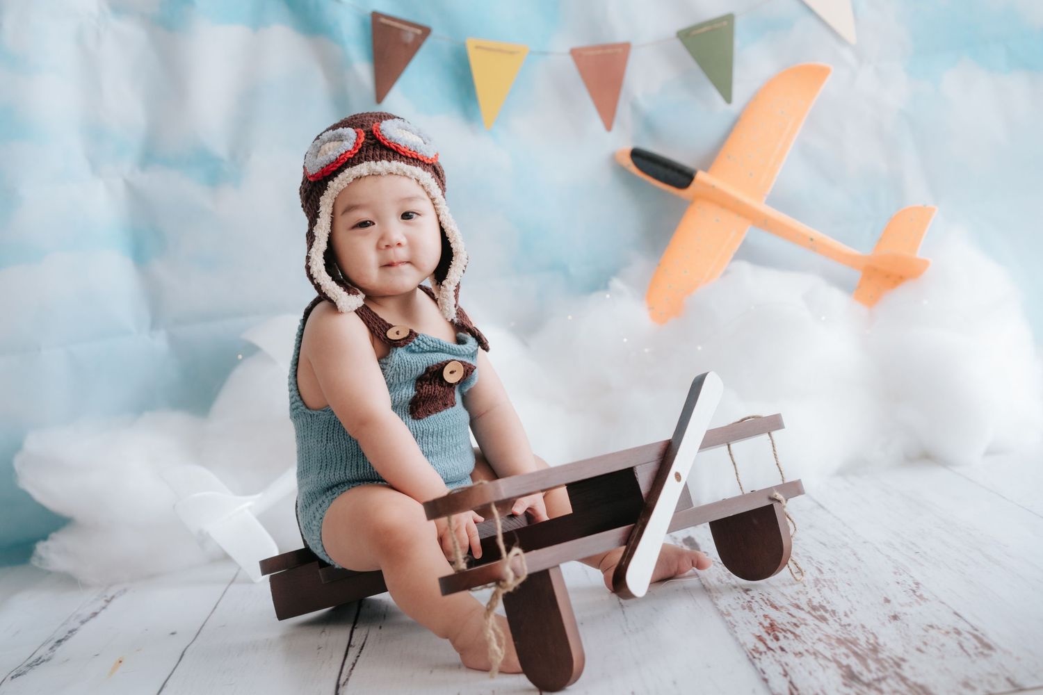 A baby sits in a white chair surrounded by floral decorations and balloons at a birthday celebration.