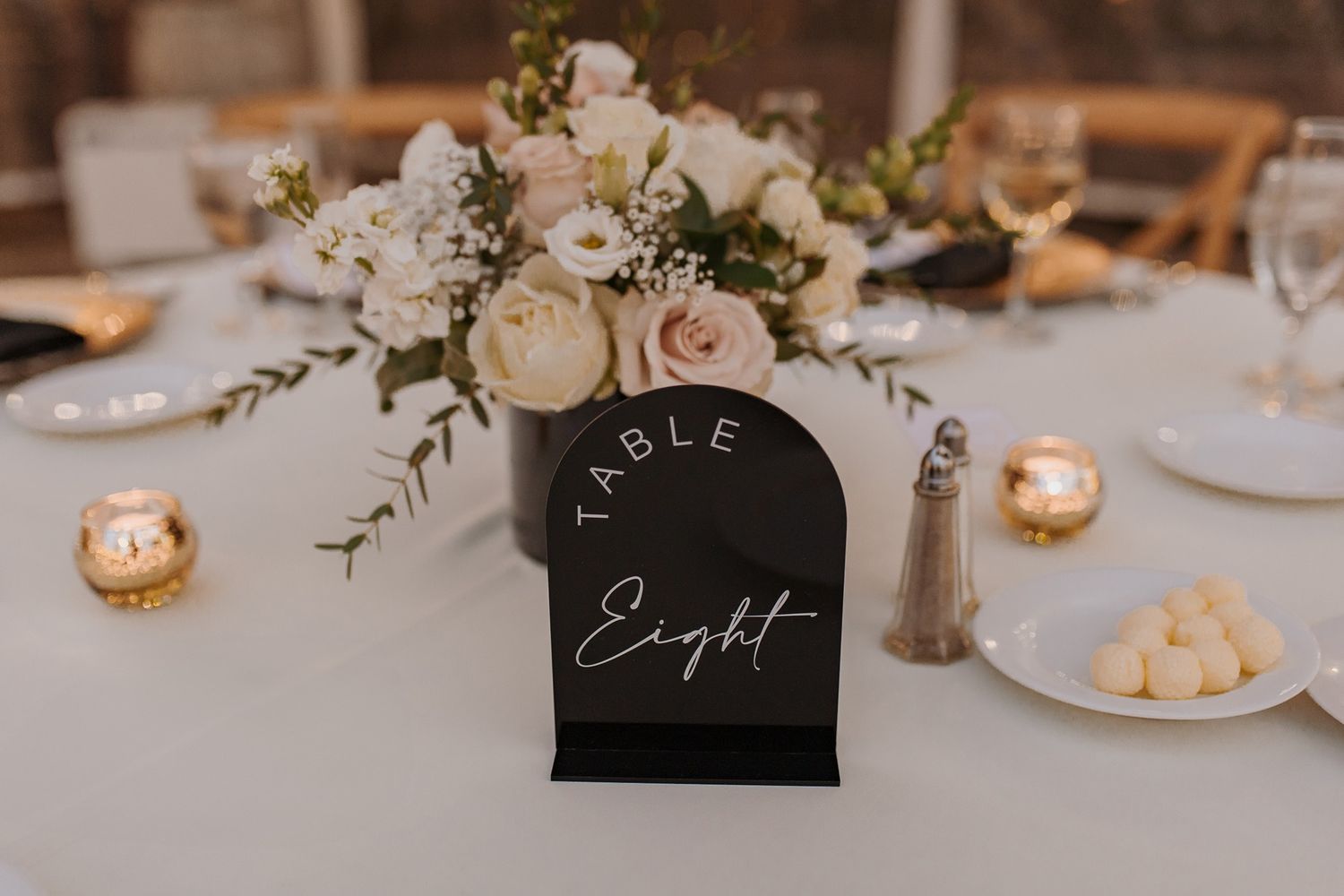 Elegant wedding reception table settings with black table numbers, white floral centerpieces, and glowing candlelight.