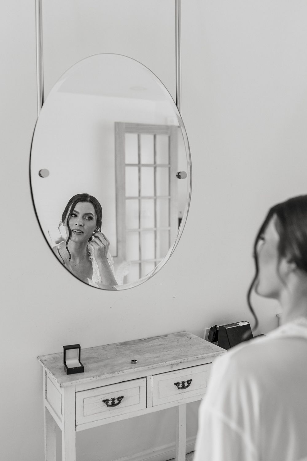 A circular mirror mounted on a white wall above a wooden dresser reflect natural light as a bride gets ready for her wedding day.