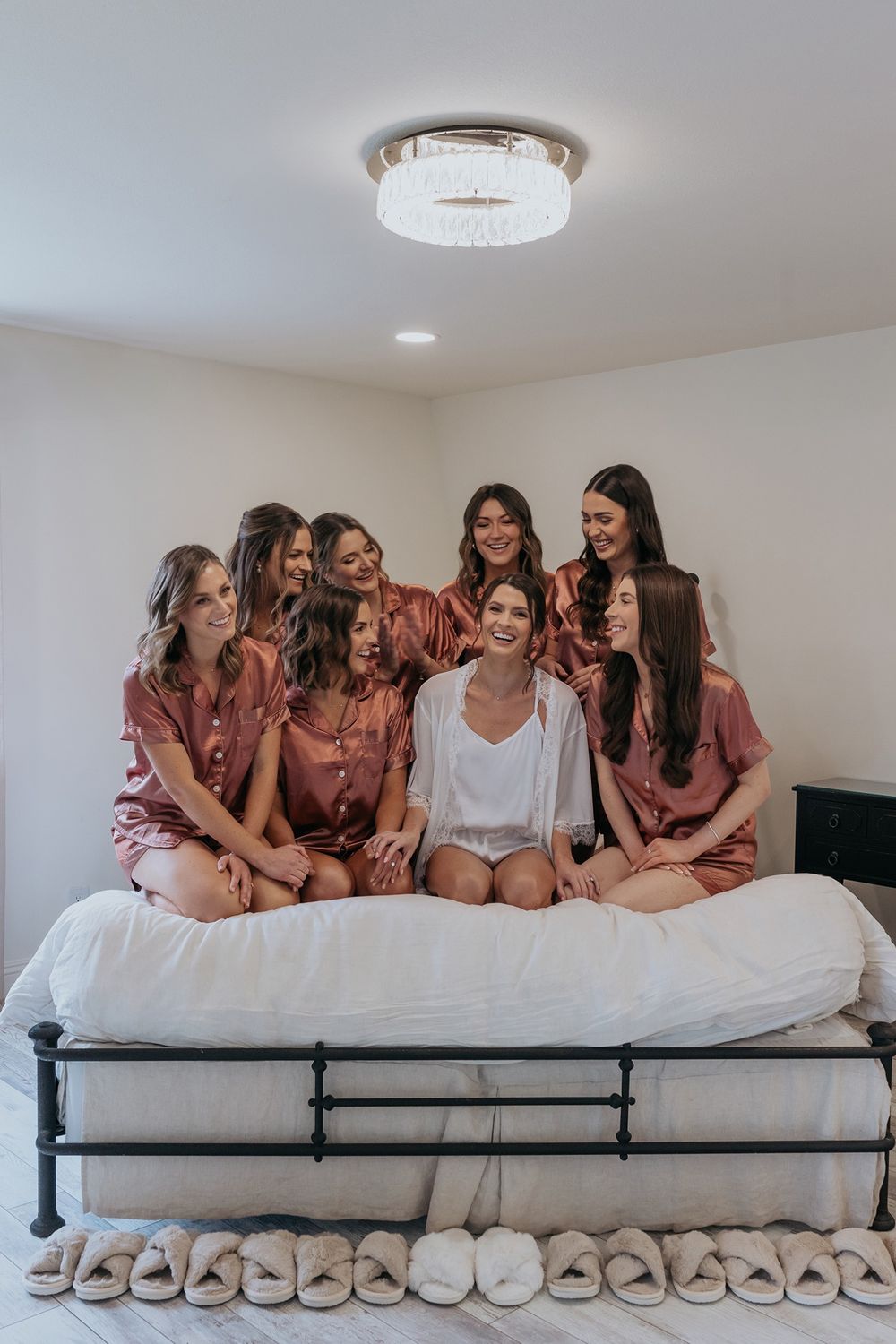 Bride and bridesmaids in matching pajamas sit together on a white bed with a row of slippers lined up on the floor at Lake Oak Meadows.