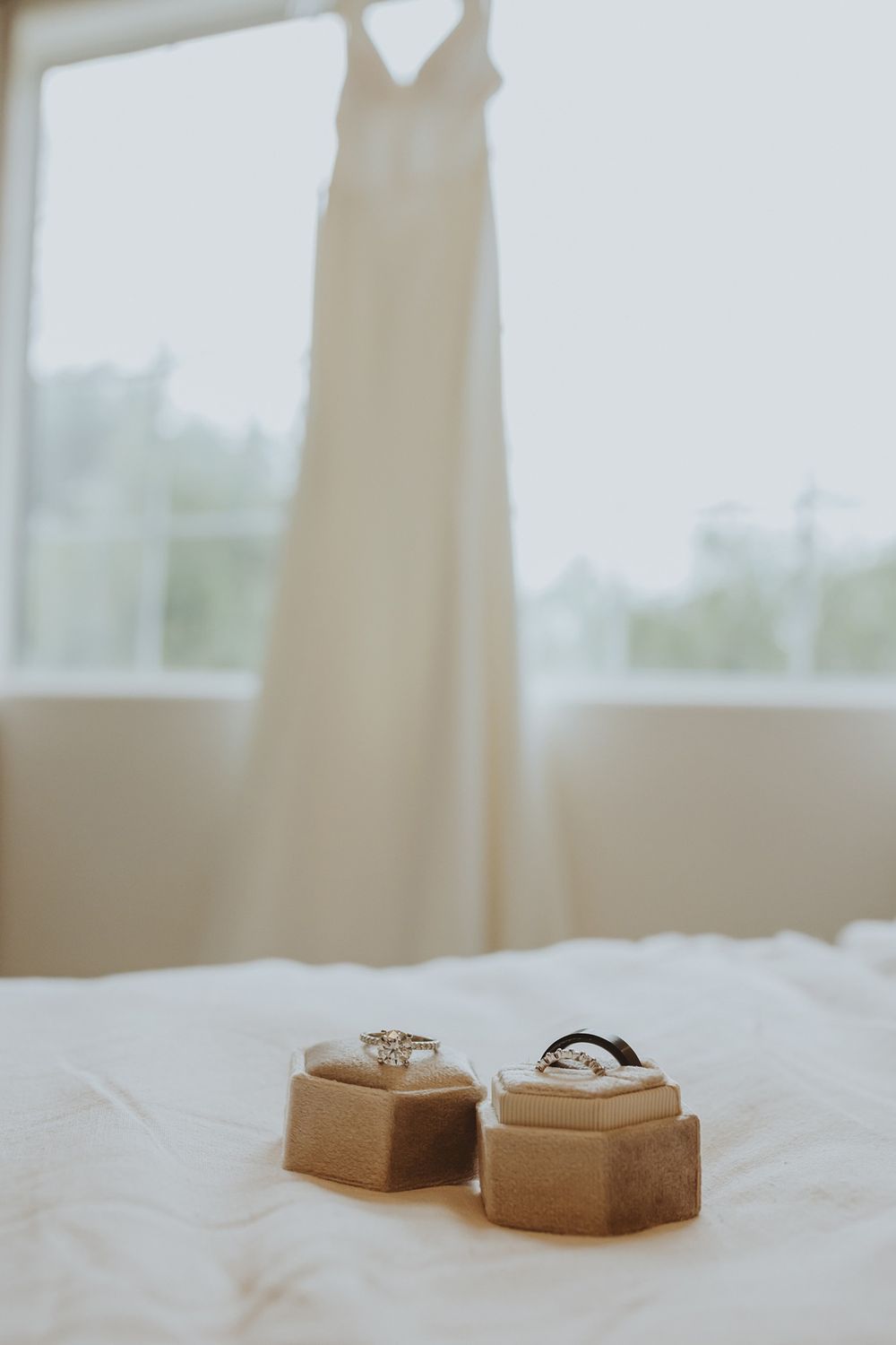 Wedding rings and jewelry displayed on neutral beige surfaces in soft natural lighting with a wedding dress hanging in the background.