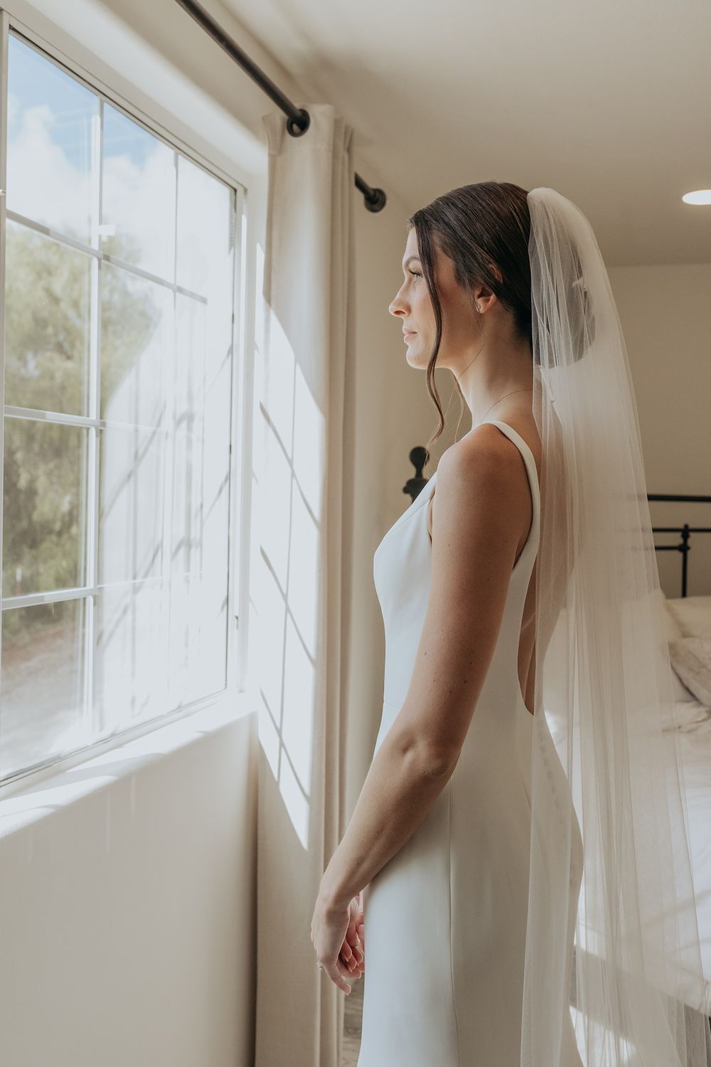 A bride in a sleek white wedding dress and veil gazes thoughtfully out a sunlit window on her special day at Lake Oak Meadows in Temecula.